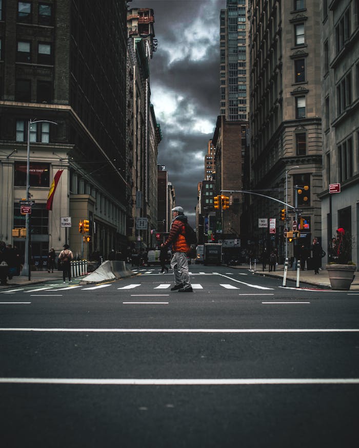 A person crossing a city street with towering skyscrapers and dramatic cloudy skies.